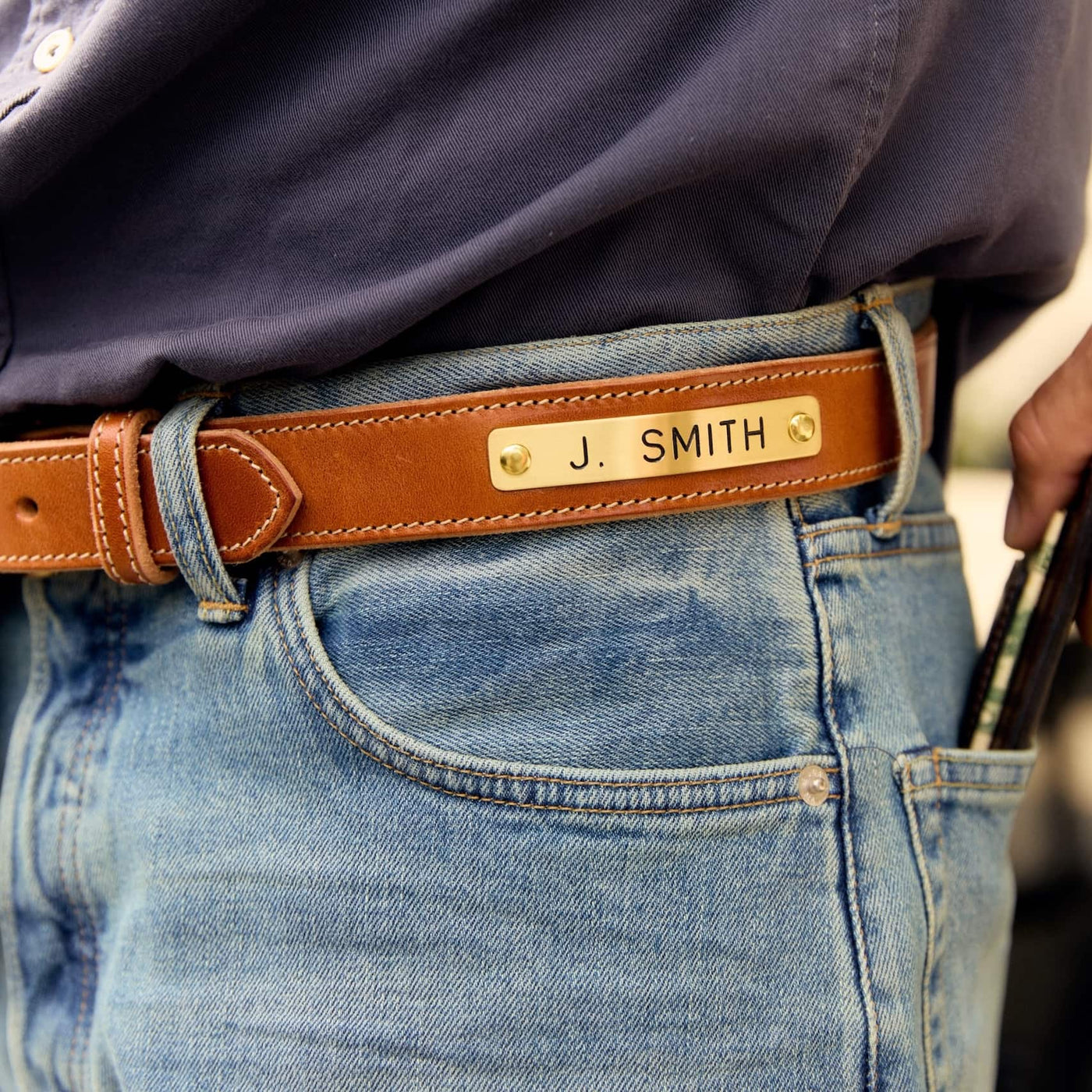 A close-up of a person wearing a tan leather belt with a gold-tone metal nameplate engraved with 'J. SMITH', paired with blue jeans and a dark navy shirt.