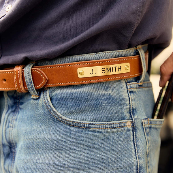 A close-up of a person wearing a tan leather belt with a gold-tone metal nameplate engraved with 'J. SMITH', paired with blue jeans and a dark navy shirt.