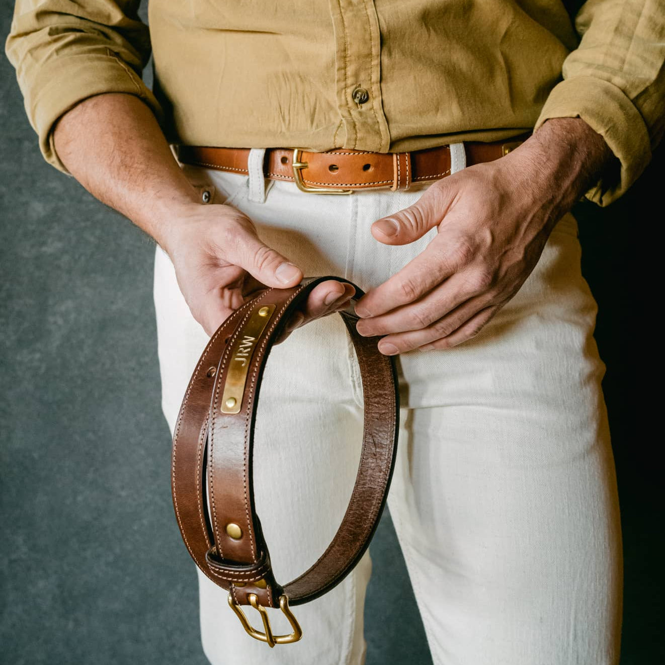 A man wearing a tan button-up shirt and white pants holds a brown leather belt with brass hardware in front of his waist, showing a close-up of the belt details.