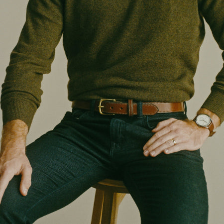 A man wearing an olive green knit sweater, a brown leather belt with a gold-tone buckle, and dark green jeans, seated on a wooden stool against a neutral beige background.